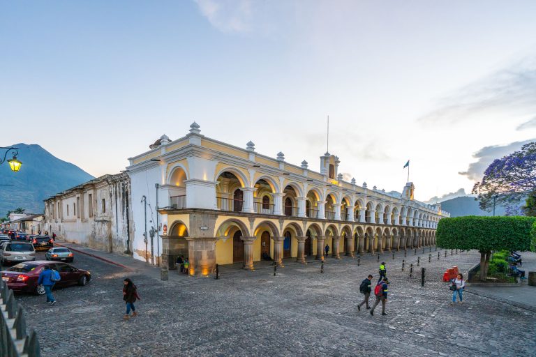 Palacio de los Capitanes Generales in Antigua Guatemala 2 Yellow and white colonial building with arched windows on a plaza with pedestrians and parked cars.