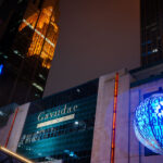 Downtown Minneapolis after dark, with the Gaviidae Common retail and office complex illuminated along Nicollet Mall. The metallic blue spherical public art installation in the foreground—inscribed with words referencing music, light, and community—adds a cultural element to the urban corridor. Reflected dramatically in the golden glass façade of the Westin building behind it is the historic Foshay Tower, one of Minneapolis’s earliest skyscrapers dating back to 1929. Today, Nicollet Mall serves as the city’s primary pedestrian and transit thoroughfare, blending modern commercial redevelopment, historic preservation, and public art into a central civic space.