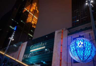 Downtown Minneapolis after dark, with the Gaviidae Common retail and office complex illuminated along Nicollet Mall. The metallic blue spherical public art installation in the foreground—inscribed with words referencing music, light, and community—adds a cultural element to the urban corridor. Reflected dramatically in the golden glass façade of the Westin building behind it is the historic Foshay Tower, one of Minneapolis’s earliest skyscrapers dating back to 1929. Today, Nicollet Mall serves as the city’s primary pedestrian and transit thoroughfare, blending modern commercial redevelopment, historic preservation, and public art into a central civic space.