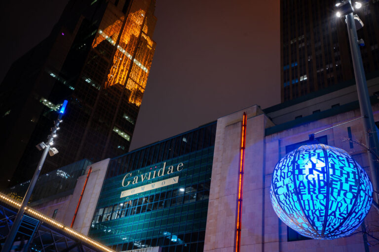 Gaviidae Common and Westin Reflection on Nicollet Mall 4 Downtown Minneapolis after dark, with the Gaviidae Common retail and office complex illuminated along Nicollet Mall. The metallic blue spherical public art installation in the foreground—inscribed with words referencing music, light, and community—adds a cultural element to the urban corridor. Reflected dramatically in the golden glass façade of the Westin building behind it is the historic Foshay Tower, one of Minneapolis’s earliest skyscrapers dating back to 1929. Today, Nicollet Mall serves as the city’s primary pedestrian and transit thoroughfare, blending modern commercial redevelopment, historic preservation, and public art into a central civic space.