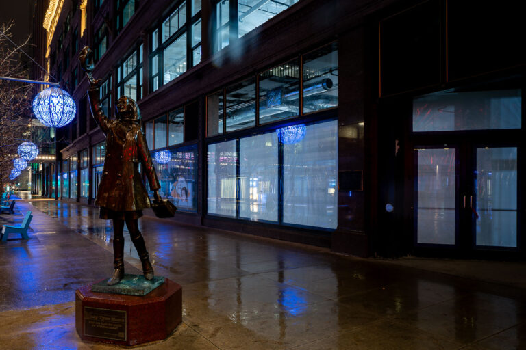 Mary Tyler Moore Statue on an Empty Nicollet Mall 2 The iconic bronze statue of Mary Tyler Moore along Nicollet Mall in downtown Minneapolis, depicting the memorable hat-tossing moment from the opening of The Mary Tyler Moore Show, which first aired in 1970 and helped cement Minneapolis in television history. Installed in 2002, the statue symbolizes the city’s cultural association with the show’s themes of independence and optimism. At the time of this photograph, Nicollet Mall—normally a busy pedestrian and transit corridor—appears unusually empty, reflecting the early days of COVID-19 shutdowns that drastically reduced public presence in city centers. Blue public art lanterns, added during street improvements completed in 2017, line the renovated streetscape, emphasizing Nicollet Mall’s evolution as both a commercial and cultural landmark.