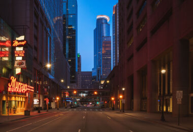A north–south view through downtown Minneapolis during early evening, with illuminated office towers forming a dense street corridor and minimal traffic on the roadway below. The cylindrical crown of the IDS Center, completed in 1973, anchors the skyline in the distance as the city’s tallest building and a defining element of its modern profile. In the foreground, the neon signage of Murray’s, a Minneapolis institution since 1946, adds a warm counterpoint to the cooler glass-and-steel architecture surrounding Nicollet Mall.