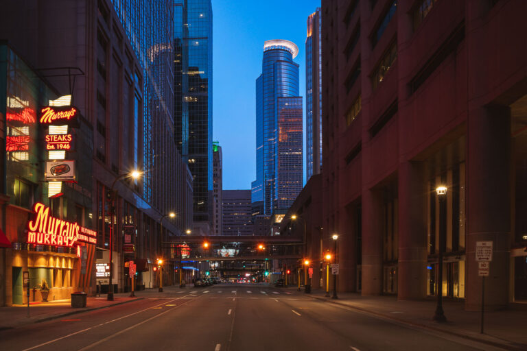 Murray's Steakhouse, Minneapolis Blue Hour 1 A north–south view through downtown Minneapolis during early evening, with illuminated office towers forming a dense street corridor and minimal traffic on the roadway below. The cylindrical crown of the IDS Center, completed in 1973, anchors the skyline in the distance as the city’s tallest building and a defining element of its modern profile. In the foreground, the neon signage of Murray’s, a Minneapolis institution since 1946, adds a warm counterpoint to the cooler glass-and-steel architecture surrounding Nicollet Mall.