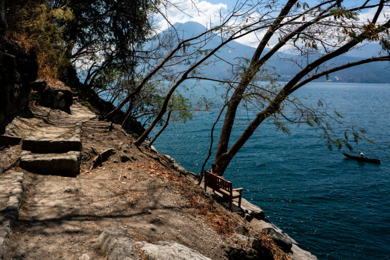 Path to Lake Atitlan, San Marcos, Guatemala 2 A dirt path with stone steps leads along the shore of Lake Atitlan near San Marcos, Guatemala, with a dugout canoe on the water and volcanoes in the distance.
