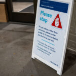 At the entrance of an Allina Health clinic, a temporary sign reminds visitors of heightened safety measures during the COVID-19 pandemic. The notice instructs patients to stop and first meet with a greeter before checking in, emphasizing that the location does not provide community COVID-19 testing and that masks may be required during the visit. These portable A-frame signs became common across medical facilities in 2020–2021, helping manage visitor flow, screen for symptoms, and reinforce evolving public health protocols at clinics and hospitals throughout Minnesota.