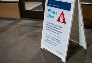 At the entrance of an Allina Health clinic, a temporary sign reminds visitors of heightened safety measures during the COVID-19 pandemic. The notice instructs patients to stop and first meet with a greeter before checking in, emphasizing that the location does not provide community COVID-19 testing and that masks may be required during the visit. These portable A-frame signs became common across medical facilities in 2020–2021, helping manage visitor flow, screen for symptoms, and reinforce evolving public health protocols at clinics and hospitals throughout Minnesota.