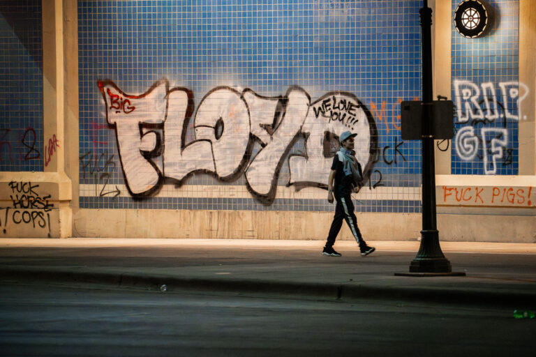 Protester walks past 'FLOYD' graffiti, Lake St, Minneapolis protests 2 A protester walks past graffiti on a tiled wall under the Hiawatha Avenue overpass near Lake Street in Minneapolis. The prominent "FLOYD" graffiti, alongside other messages like "RIP GF," emerged during the second day of protests in late May 2020. These protests erupted across Minneapolis and subsequently worldwide, following the death of George Floyd while in police custody. The messages on the infrastructure served as a public memorial and a direct expression of community outrage against systemic racism and police brutality.