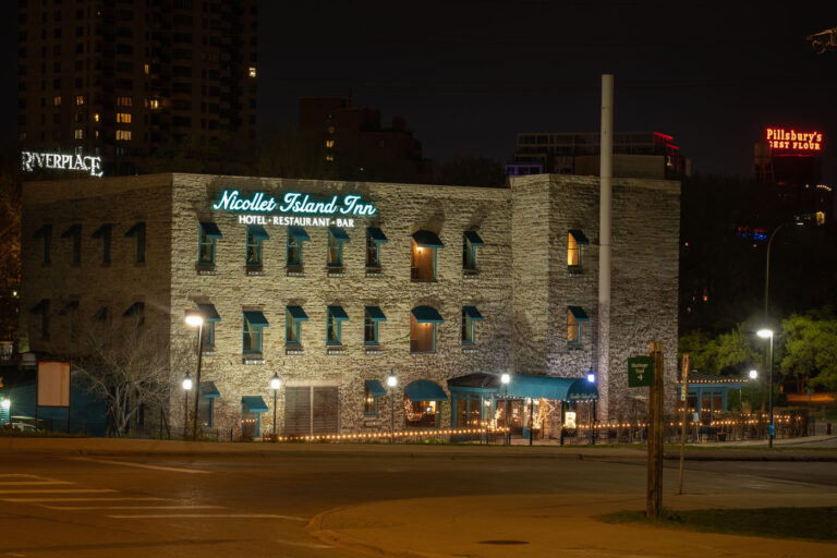 Nicollet Island Inn at Night, Minneapolis Riverfront 3 The Nicollet Island Inn, a landmark along the Mississippi River in Minneapolis, occupies a 19th-century brick and limestone structure originally built in the 1890s as an industrial facility. Converted into a hotel and restaurant in the 1970s, it retains its historic character with exposed stone walls and vintage architectural details. Illuminated signage and soft outdoor lighting highlight the inn’s distinctive facade, with the iconic Pillsbury’s Best Flour sign visible across the river, linking the scene to the city’s milling heritage.