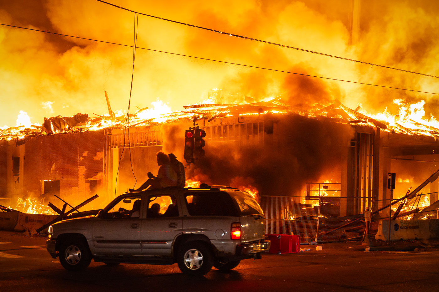 People on the roof of a car as a large building burns behind them