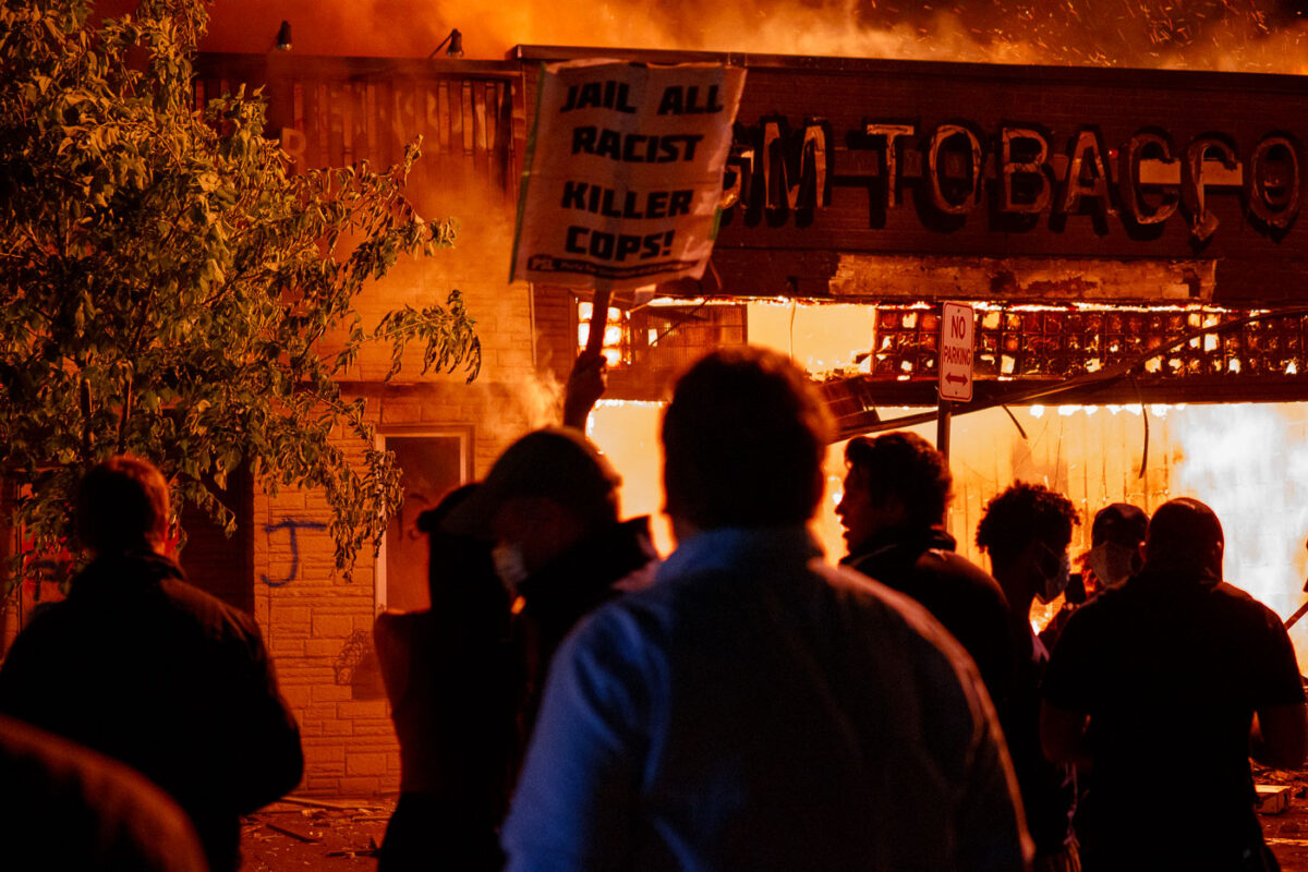 A protester holds up a sign reading "Jail all racist killer cops!" outside a burning GM Tobacco store on East Lake Street on May 28th, 2020 during the 3rd day of protests in Minneapolis following the death of George Floyd.