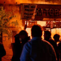 A protester holds up a sign reading "Jail all racist killer cops!" outside a burning GM Tobacco store on East Lake Street on May 28th, 2020 during the 3rd day of protests in Minneapolis following the death of George Floyd.