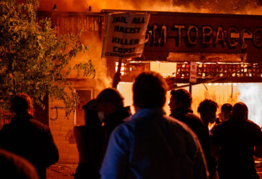 A protester holds up a sign reading "Jail all racist killer cops!" outside a burning GM Tobacco store on East Lake Street on May 28th, 2020 during the 3rd day of protests in Minneapolis following the death of George Floyd.