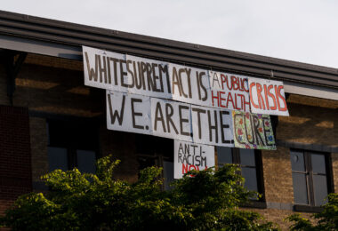 A sign reading "White supremacy is a public health crisis. We.Are.The.Cure. Anti-Racism NOW" hanging over the side of the Minneapolis Police Third Precinct on May 31, 2020.