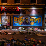A large George Floyd mural on 38th Street with flowers left behind in front of it. George Floyd was killed around the corner on Chicago Ave the month prior.