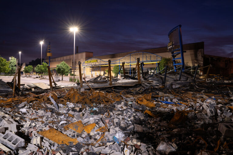 Burned Dollar General & HR Block, Hi-Lake Shopping Center 1 The Hi-Lake Shopping Center on East Lake Street in South Minneapolis shows extensive fire damage. In the foreground are the charred remains of an HR Block store, while a Dollar General store in the background exhibits significant structural damage and boarded windows. This commercial property sustained widespread destruction in May 2020, following the murder of George Floyd. The damage to businesses along Lake Street became a notable aspect of the protests and civil unrest that impacted the city, affecting local services and the community.