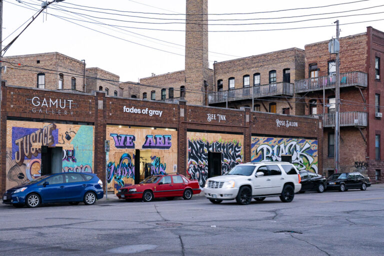 Gamut Gallery boarded storefronts with murals, Minneapolis 1 Storefronts along a street in Minneapolis, including Gamut Gallery, are boarded with plywood. These boards were installed to protect businesses during the civil unrest and protests that followed the murder of George Floyd in May 2020. Many of the plywood panels were subsequently transformed into murals and public art, reflecting themes of justice and community. Businesses like Gamut Gallery, 'fades of gray,' 'BEE INK,' and 'ROSE SALON' participated in this widespread practice across the city.