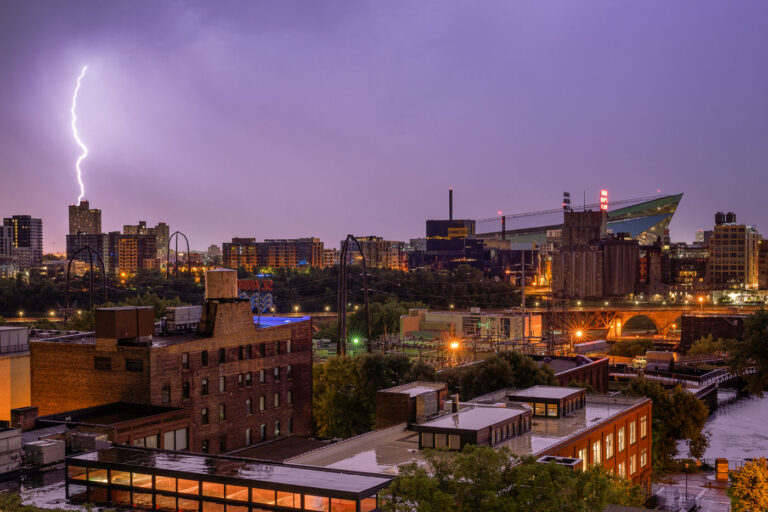A lightning bolt illuminates the early morning sky over downtown Minneapolis on July 18, 2020, striking a high-rise building. In the background, US Bank Stadium, the home of the Minnesota Vikings since its opening in 2016, stands prominently with its distinctive angular roof. The surrounding urban environment includes various commercial and residential structures, alongside industrial remnants and infrastructure near the Mississippi River. This event occurred during a period of significant social and political activity in Minneapolis.