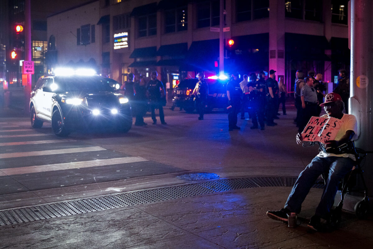 A homeless man sits on Nicollet Mall asking for help while riot police move in after rioting broke out following the false rumors of a downtown shooting by the police. The man died of a suicide.