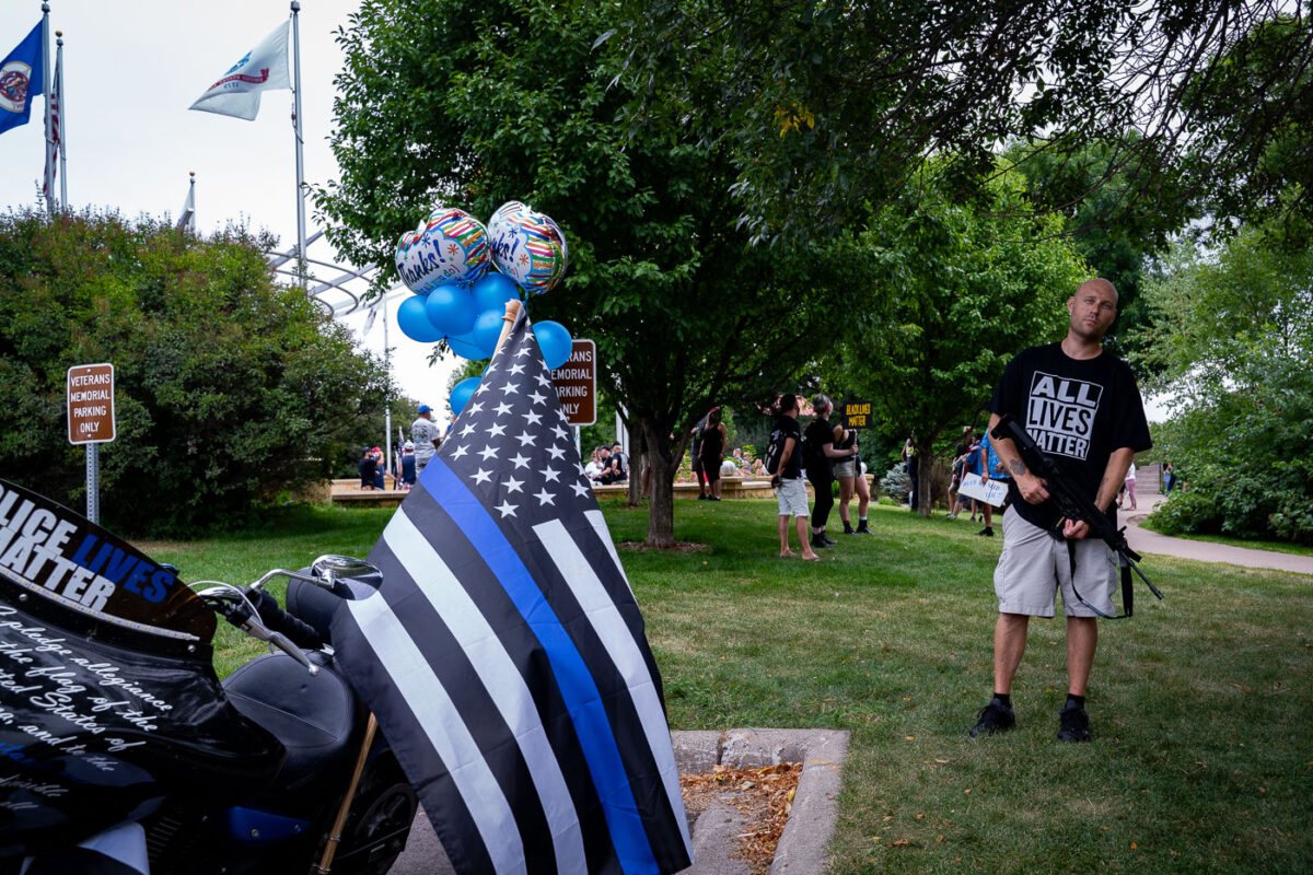 Man stands with a large gun with Black Lives Matters protesters behind him at a  "Back The Blue" rally in Woodbury, Minnesota on August 8th, 2020.