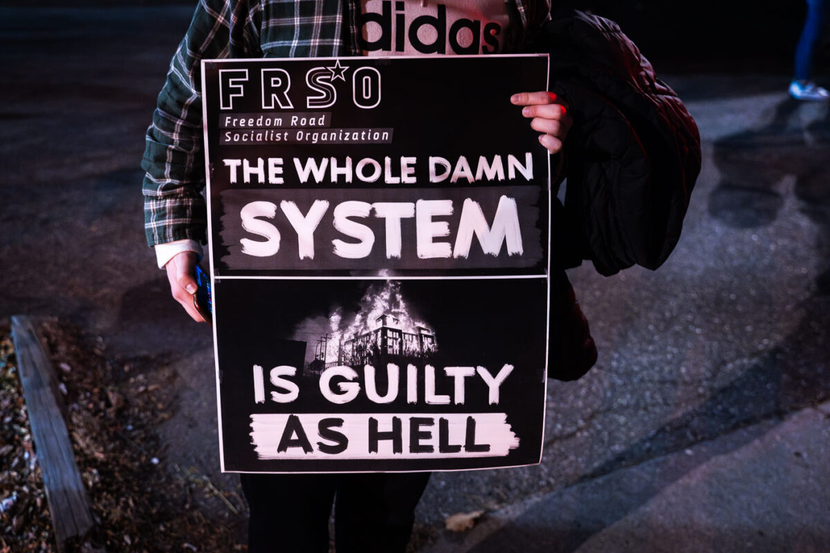 A protester holds up a sign by the Freedom Road Socialist Organization (FRSO). It reads “The whole damn system is guilty as hell”.