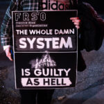 A protester holds up a sign by the Freedom Road Socialist Organization (FRSO). It reads “The whole damn system is guilty as hell”.