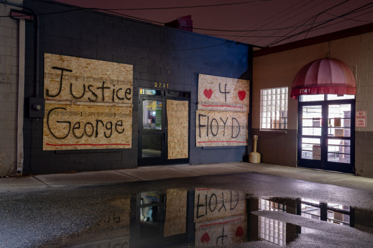 Minneapolis Uprising: Justice 4 George Floyd Boards, Hennepin Ave 1 Plywood boards bearing the message 'Justice 4 George Floyd' cover the windows of a retail store at 2741 Hennepin Avenue in Uptown Minneapolis. These boards were installed during the Minneapolis Uprising in May and June 2020, a period of widespread protest and civil unrest following the murder of George Floyd by Minneapolis police. Many businesses in Minneapolis boarded up their windows to protect against potential damage, and these surfaces often became canvases for public art and messages related to the movement. The graffiti art on these boards served as a visual testament to the community's demand for justice and accountability, reflecting the widespread impact of the uprising on the urban environment.