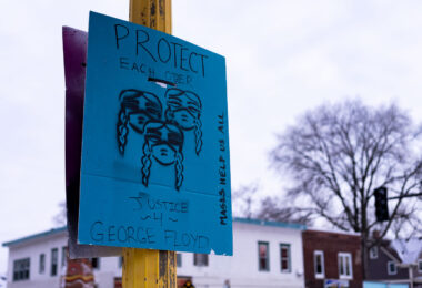 A sign at George Floyd Square that reads "Protect Each Other" "Justice 4 George Floyd" "Masks Help Us All"