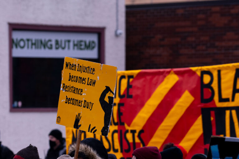 St. Paul Counter Protest: "When Injustice Becomes Law" Sign 2 A protester holds a sign reading "When Injustice becomes Law Resistance becomes Duty" during a march in St. Paul, Minnesota. The sign's message reflects a sentiment of civil disobedience in response to perceived legal injustices. In the background, a banner displays "BLACK LIVES MATTER" and slogans such as "COMMUNITY NOT COPS" and "PEOPLE NOT PIPELINES," indicating the protest's alignment with broader social justice movements. The presence of these messages suggests the event was a counter-protest or a demonstration related to the Minneapolis Uprising and ongoing calls for police reform and racial equity.