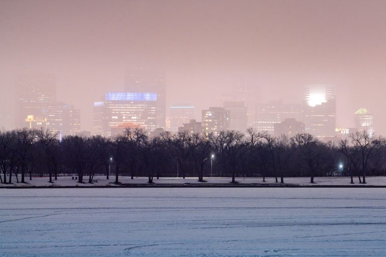 Downtown Minneapolis skyline rises above bare trees and a frozen lake shrouded in winter fog.