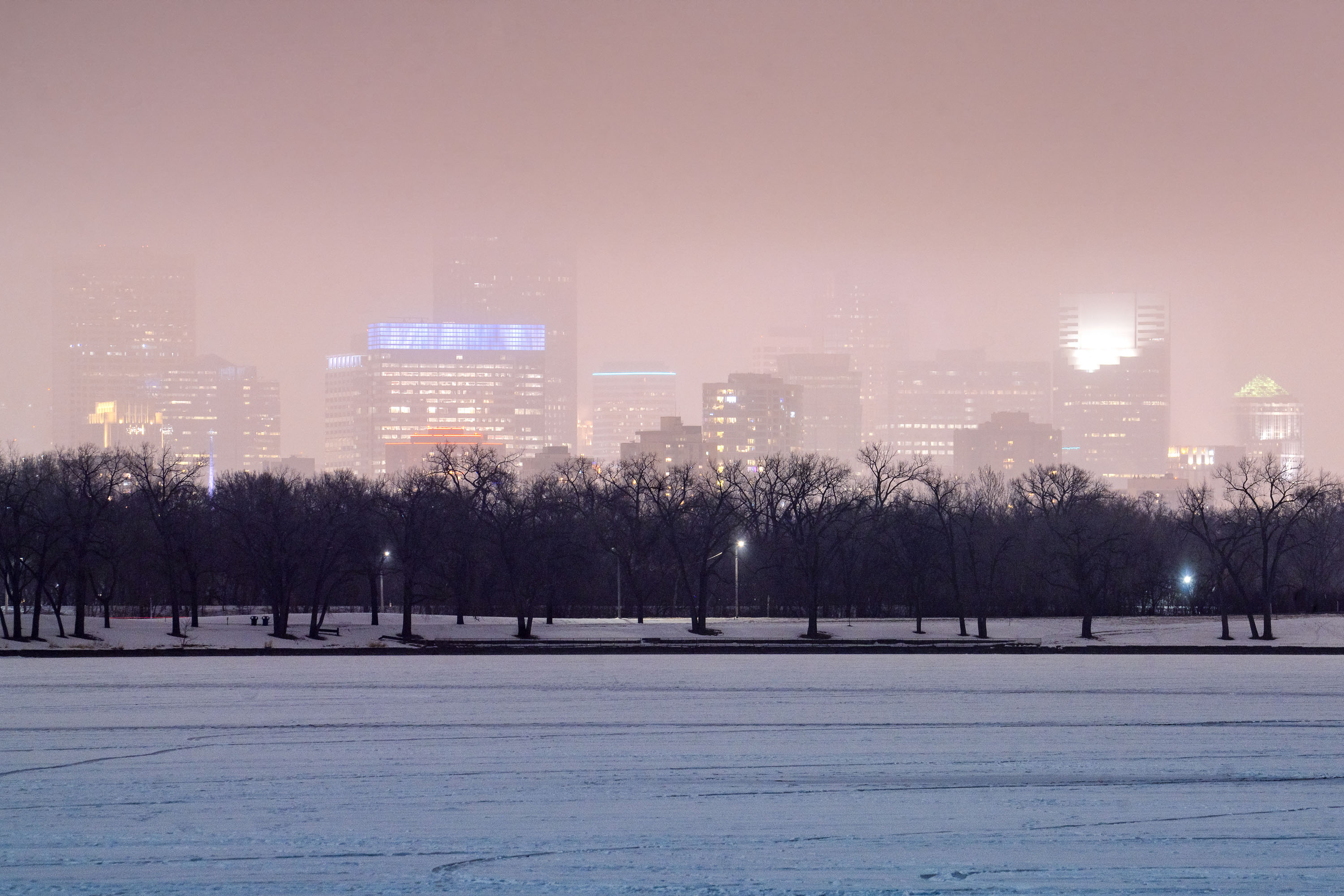 Downtown Minneapolis skyline rises above bare trees and a frozen lake shrouded in winter fog.