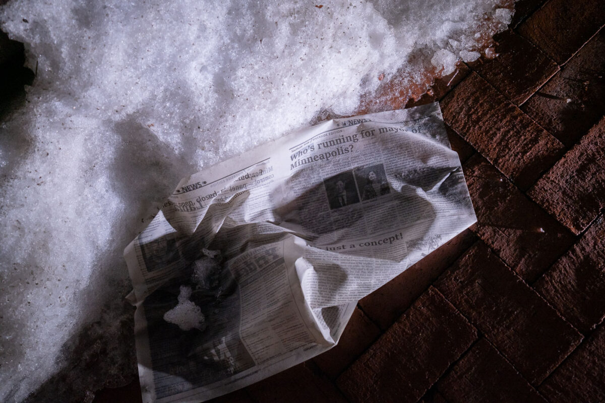 A wet newspaper lays in the snow outside the Hennepin County Government Center. Article titled “Who’s running for Mayor of Minneapolis?”