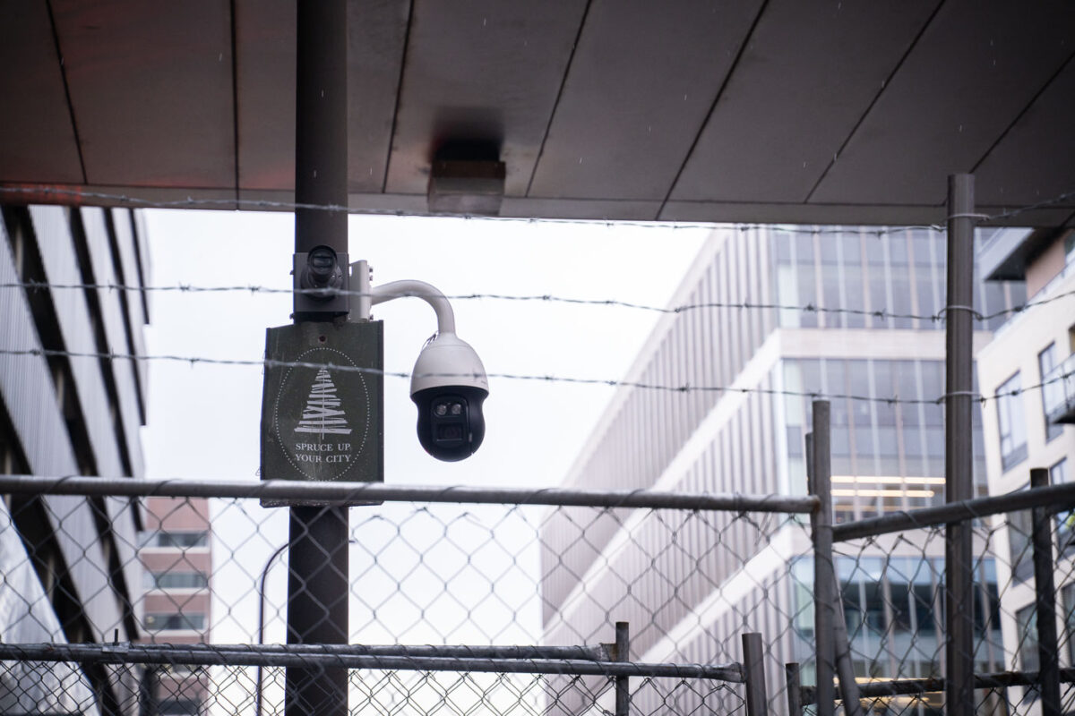 A sign reading "Spruce Up Your City" next to a surveillance camera behind barbed wire in downtown Minneapolis.