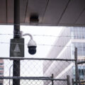 A sign reading "Spruce Up Your City" next to a surveillance camera behind barbed wire in downtown Minneapolis.