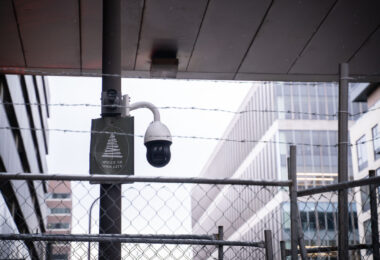 A sign reading "Spruce Up Your City" next to a surveillance camera behind barbed wire in downtown Minneapolis.