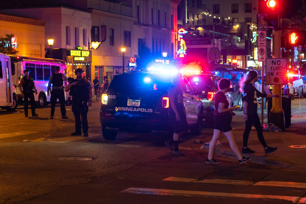 Bar patrons walk down Hennepin Avenue past armed Minneapolis police who responded to those protesting the June 3rd shooting death by a Federal task force.