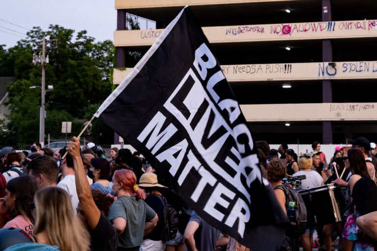 Black Lives Matter sign and protesters in Uptown Minneapolis 1 Protester holds up Black Lives Matter flag while marching down Lake Street during a march following the June 3rd and June 13th deaths of Winston Smith and Deona Marie.Winston Smith was killed after Hennepin and Ramsey County officers fired their weapons while part of a Federal Task Force serving a warrant. Deona Marie was killed when Nicholas Kraus drove his vehicle into those protesting the killing of Smith.