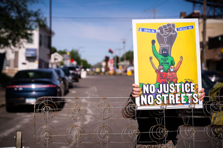 Protester holds up sign at George Floyd Square street barricade 3 A woman holds a "No Justice No Streets" yard sign on the day the city tried to clear out barricades around the square.
