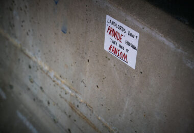 A handwritten sticker criticizing property ownership dynamics is affixed to a concrete wall in downtown Minneapolis. Reading “Landlords don’t ‘provide’ housing, they hold it ransom,” the message reflects growing public frustration with rising rents and the commodification of housing. Such small-scale urban protest art has become a recurring visual language across Minneapolis since the 2020s, echoing broader debates about housing justice and economic inequality.