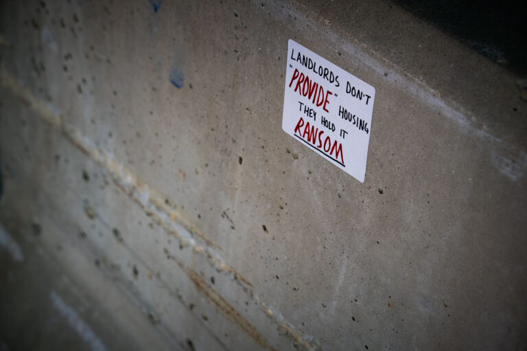 Anti-Landlord Sticker on Downtown Wall — Minneapolis 3 A handwritten sticker criticizing property ownership dynamics is affixed to a concrete wall in downtown Minneapolis. Reading “Landlords don’t ‘provide’ housing, they hold it ransom,” the message reflects growing public frustration with rising rents and the commodification of housing. Such small-scale urban protest art has become a recurring visual language across Minneapolis since the 2020s, echoing broader debates about housing justice and economic inequality.
