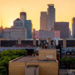 The downtown Minneapolis skyline glows at sunset, viewed from the south side near the Elliot Park and Stevens Square neighborhoods. Prominent landmarks include the IDS Center, Capella Tower, and Wells Fargo Center—each a hallmark of the city’s late-20th-century architectural boom. The mix of older mid-rise apartment structures and new glass towers highlights the city’s ongoing evolution, where historic housing blocks now share the horizon with symbols of modern corporate development.