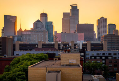 The downtown Minneapolis skyline glows at sunset, viewed from the south side near the Elliot Park and Stevens Square neighborhoods. Prominent landmarks include the IDS Center, Capella Tower, and Wells Fargo Center—each a hallmark of the city’s late-20th-century architectural boom. The mix of older mid-rise apartment structures and new glass towers highlights the city’s ongoing evolution, where historic housing blocks now share the horizon with symbols of modern corporate development.