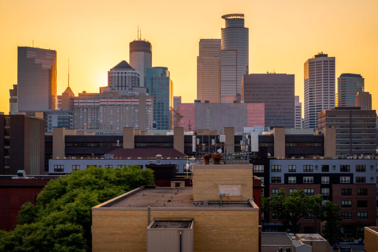 Minneapolis Skyline at Sunset from the South Loop 3 The downtown Minneapolis skyline glows at sunset, viewed from the south side near the Elliot Park and Stevens Square neighborhoods. Prominent landmarks include the IDS Center, Capella Tower, and Wells Fargo Center—each a hallmark of the city’s late-20th-century architectural boom. The mix of older mid-rise apartment structures and new glass towers highlights the city’s ongoing evolution, where historic housing blocks now share the horizon with symbols of modern corporate development.