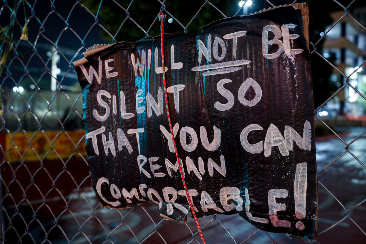 A sign reading "We will not be silent so that you can remain comfortable!" on the security fencing around the Winston Smith and Deona Marie Memorials at Lake & Girard.