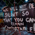 A sign reading "We will not be silent so that you can remain comfortable!" on the security fencing around the Winston Smith and Deona Marie Memorials at Lake & Girard.