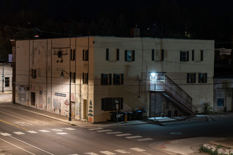 Cloquet, MN: Building with murals at night 2 A building in Cloquet, Minnesota, features exterior murals of storefronts and Parisian scenes at night. An external staircase leads to an upper-floor entrance.