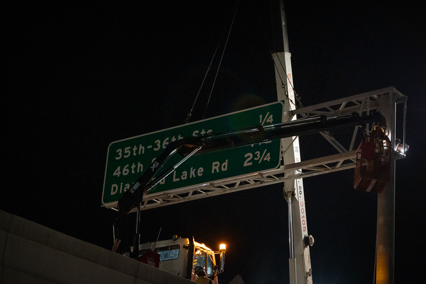 A crane installs a new directional sign for 35th-36th St, 46th Ave, and Diamond Lake Rd at night, guiding traffic in an urban environment.