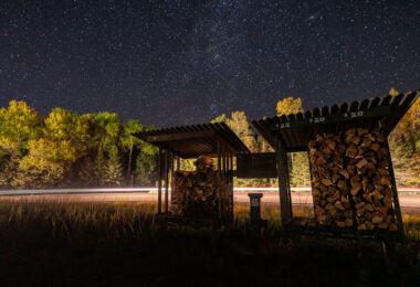 A small roadside firewood stand sits quietly beneath a star-filled sky near Isabella in northern Minnesota, a gateway community to the Superior National Forest. These honor-system stands are common throughout the Northwoods, supplying bundled campfire wood to travelers headed toward nearby campgrounds, trailheads, and remote lake cabins. Long-exposure headlights from passing vehicles streak across the background, contrasting with the stillness of the surrounding forest and the clear autumn night above.