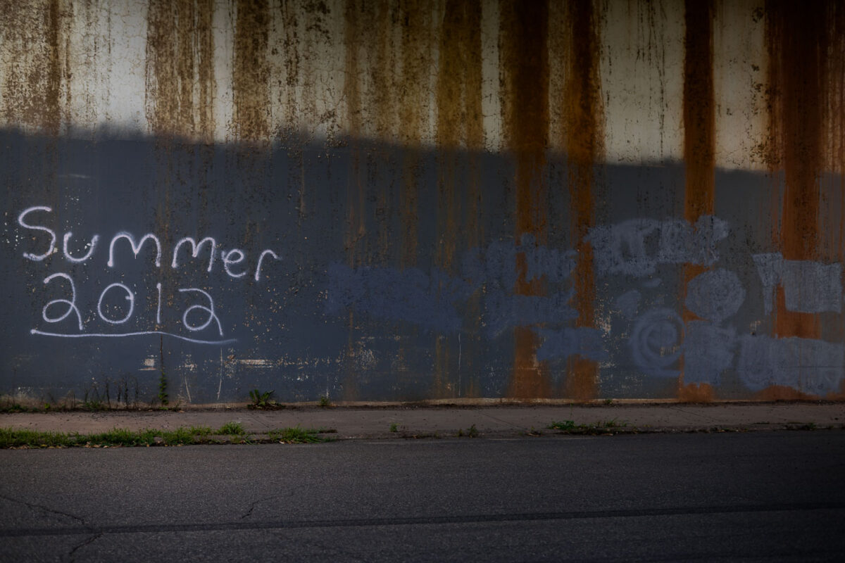 Graffiti reading "Summer 2012" spray-painted on a rusted, weathered metal wall, likely part of an industrial or infrastructure site.