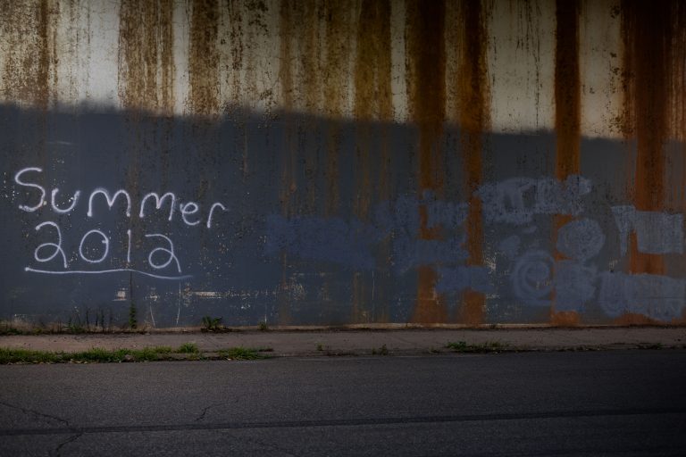 Summer 2012 Graffiti on Rusted Metal Wall 1 Graffiti reading “Summer 2012” on a rusted metal wall beside a sidewalk.