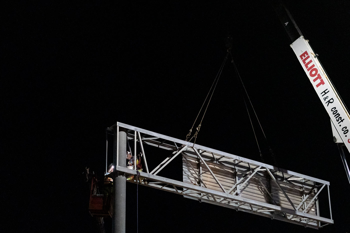 Workers install a large overhead sign structure at night using a crane, essential for highway traffic management.